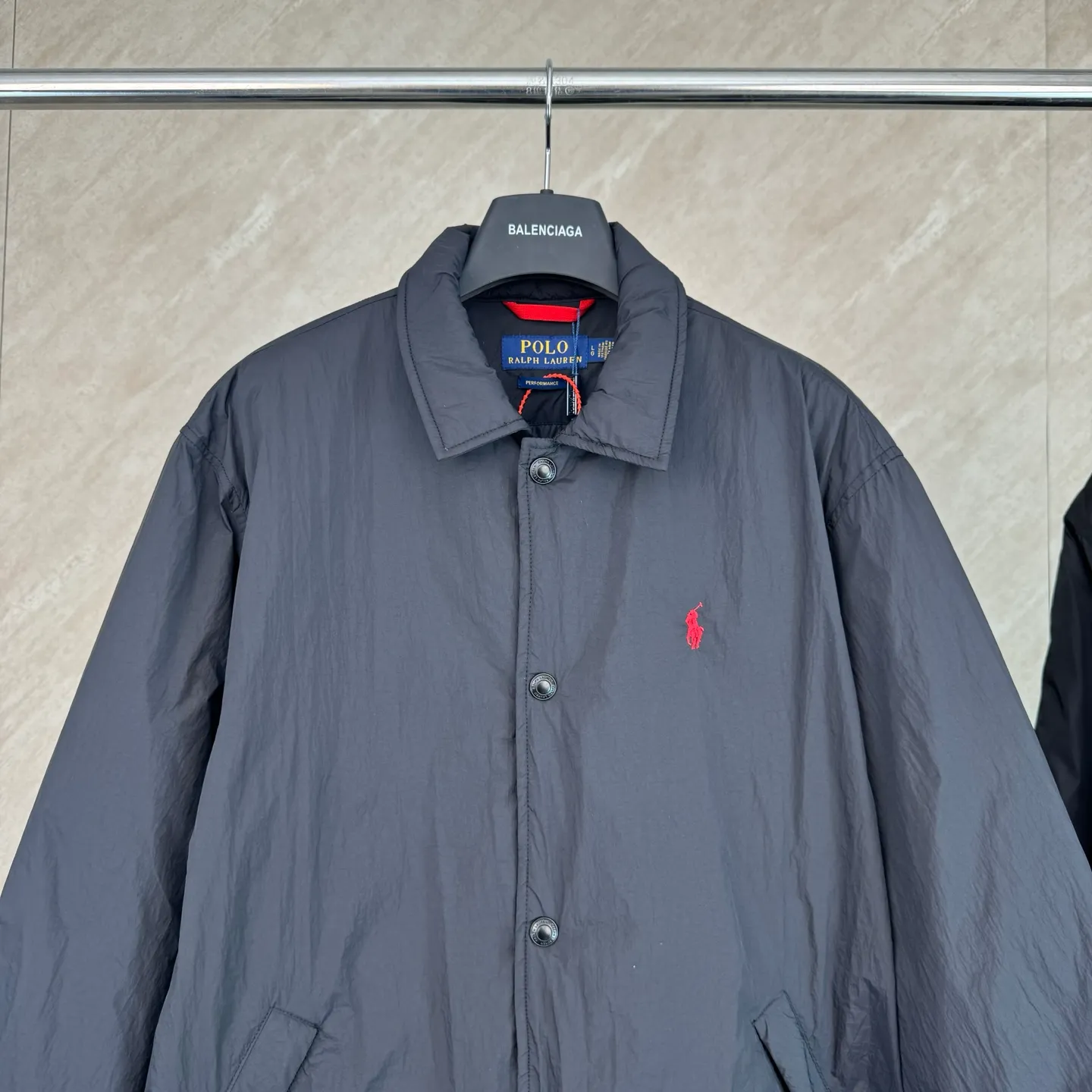 P670 25FW Ralph Lauren red label light cotton jacket (coach jacket) size s m l xl Fabric procurement: a combination of pragmatism and classic texture Material: high-density nylon taffeta Features: density is about 210T-300T, smooth and slightly matte surface, excellent windproof and waterproof performance (with DWR water-repellent treatment), strong wrinkle resistance and easy care, in line with the positioning of American casual "durable and practical".  Color: Classic dark black, using vat dyeing process (dye penetrates into the fiber), the color fastness reaches ISO level 4 or above to avoid fading or color transfer after washing.  2. Lining fabric Material: Lightweight cotton-polyester blended lining (65% Polyester + 35% Cotton) Function: Reduce friction between the fabric and inner clothing, improve wearing smoothness; blended material has both breathability and durability to avoid the stuffy feeling of pure nylon lining.  Version and cutting process Version: Classic Coach Jacket, loose version, slightly dropped shoulder line (shoulder width is 1-2cm wider than the standard version), length to the hips, and slightly tapered straight sleeves, taking into account both freedom of movement and casual style.  Embroidery technology: Polo horse logo on the left chest: flat satin stitch embroidery (satin embroidery), embroidery thread: 100% polyester high color fastness embroidery thread (the color number is consistent with the brand’s standard red color card), stitch density 12-15 stitches/cm, the outline of the horse logo is clear, and the details of the rider and horse are burr-free.  Embroidery positioning: precise positioning through the template (12cm from the neckline and 8cm from the placket) to ensure left and right symmetry and centering.  Topstitching technology: The seams of the body are made of double-needle flat seams (needle pitch 3cm/10 needles), the stitches are smooth and straight, the stitching color is dark black (the same color as the fabric), and the stitch width is 0.5cm, which enhances firmness while forming simple decorative lines.  1. Main body splicing, splicing of the front placket and body: The placket is made of double-layer folding + flat seam splicing, with a thickness of about 0.3cm to ensure that the buttons are not easily deformed when fixed; the edges of the placket are cut with a hot knife to avoid burrs.  Sleeve panels are spliced ​​to the body: the armholes are spliced ​​with round-head keyhole machine seams + flat seams. After splicing, the armholes have a natural curvature and no sense of tightness during movement; the height of the sleeves is about 8-10cm, which is in line with the loose characteristics of casual jackets.  Main button: black urea resin button, matte surface treatment, buttonholes are hand-made keyholes (6 strands of cotton thread, keyhole density 8 stitches/mm), the edges of the eyelets are smooth and without burrs.  Collar buttons: The first button on the collar is a hidden button (diameter 12mm), which prevents the collar from opening and improves the neatness of wearing.